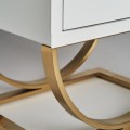 Moderní bílý noční stolek Bynum ve skleněném provedení se dvěma šuplíky a zaoblenou kovovou podstavou ve zlaté barvě 60cm