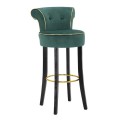 Designová glamour kulatá barová židle Vivienne se smaragdově zeleným semišovým potahem se zlatým lemováním s dekorativními knoflíčky v zámeckém stylu s černými hranatými nožičkami s kovovou kulatou opěrkou na nohy ve zlaté barvě